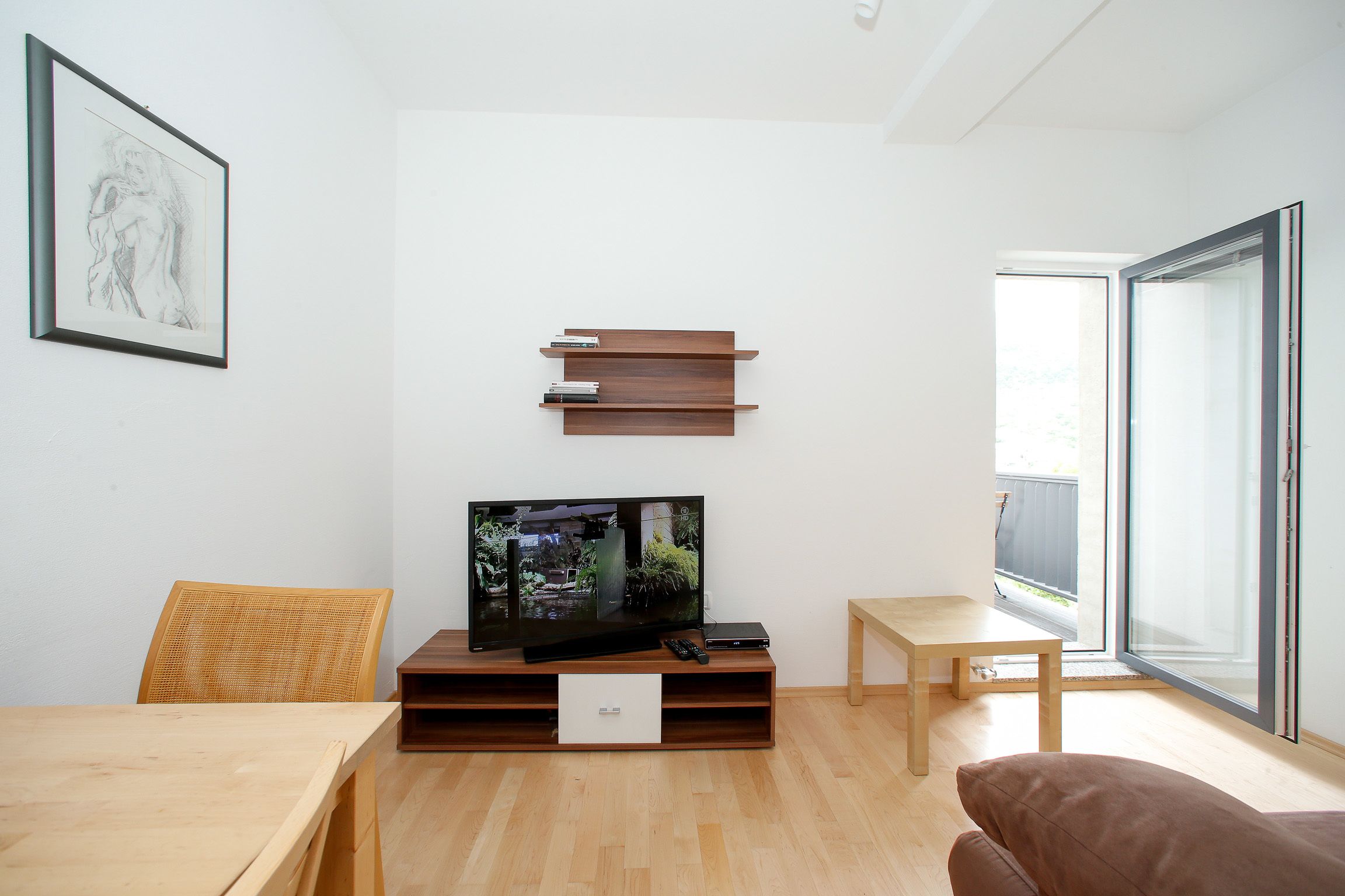 Uma sala de estar moderna com televisão de tela plana sobre suporte de madeira, uma mesa de jantar, um sofá e uma grande janela que oferece luz natural.