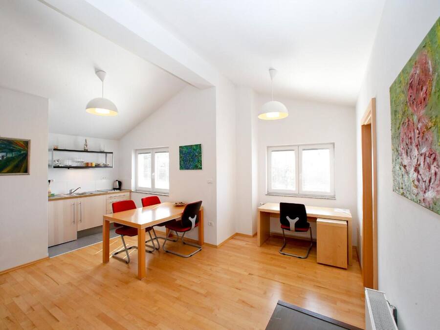 A modern kitchen with white cabinets, a dining area with a wooden table and red chairs, and a workspace with a desk and chair.