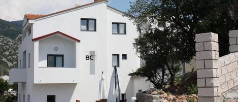 A two-story white building with a red roof and a small balcony. There are two windows on the upper floor and one on the lower floor. A sign with the letters "BC" is visible on the building.