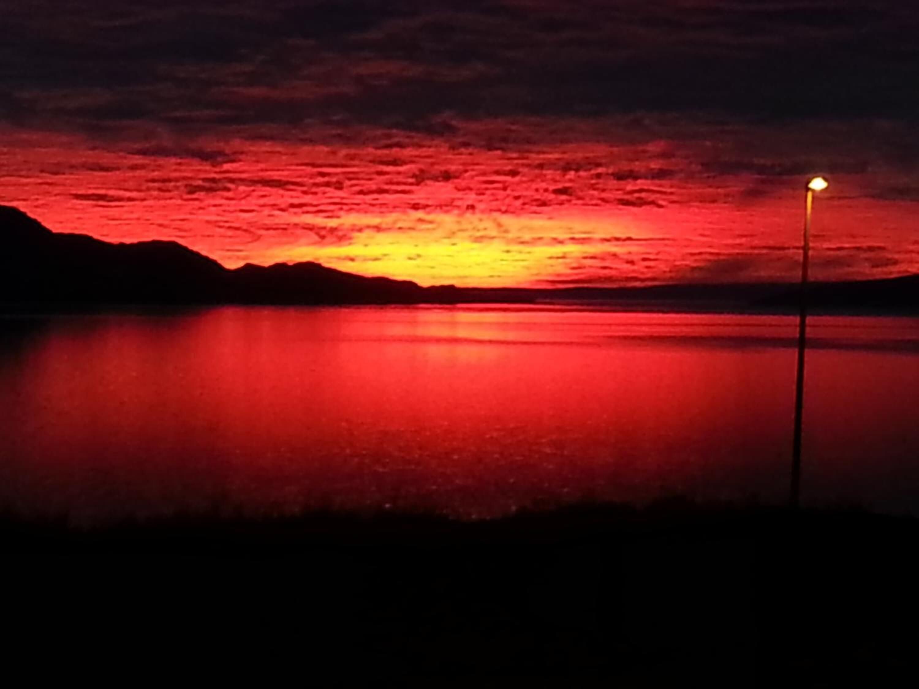 A sunset over a calm body of water with a silhouette of mountains in the background and a single light post in the foreground.