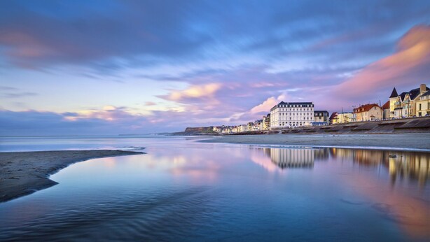 Un hôtel situé sur la côte, avec une étendue d’eau calme en premier plan, une rangée de bâtiments le long de la plage et un ciel dégagé.
