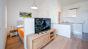 Un appartement moderne comprenant un meuble de rangement en bois pour l’équipement audiovisuel, un lit, un téléviseur et une kitchenette.