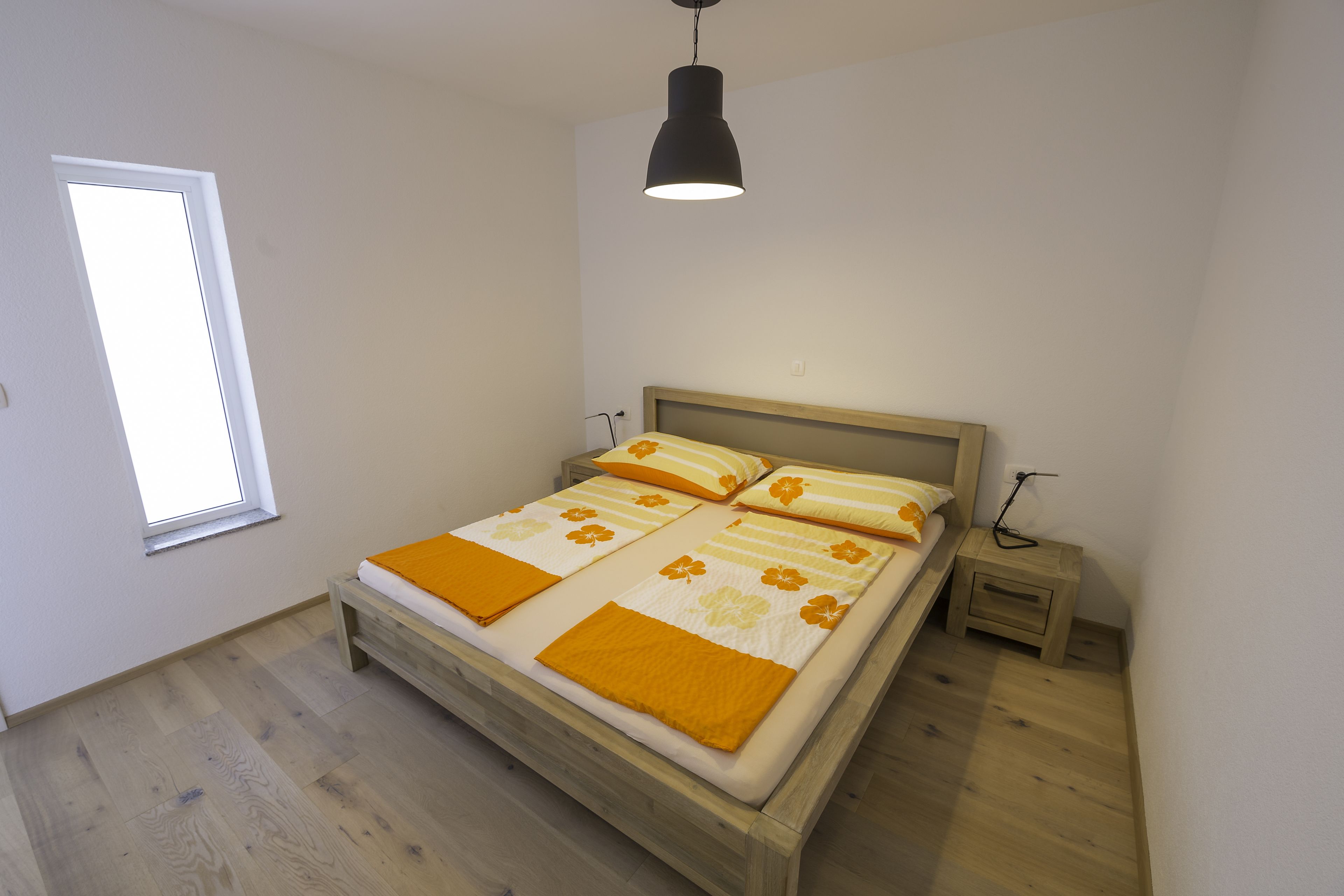 A bedroom with a wooden bed, a bedside table, a nightstand, a window, and a hanging light fixture.
