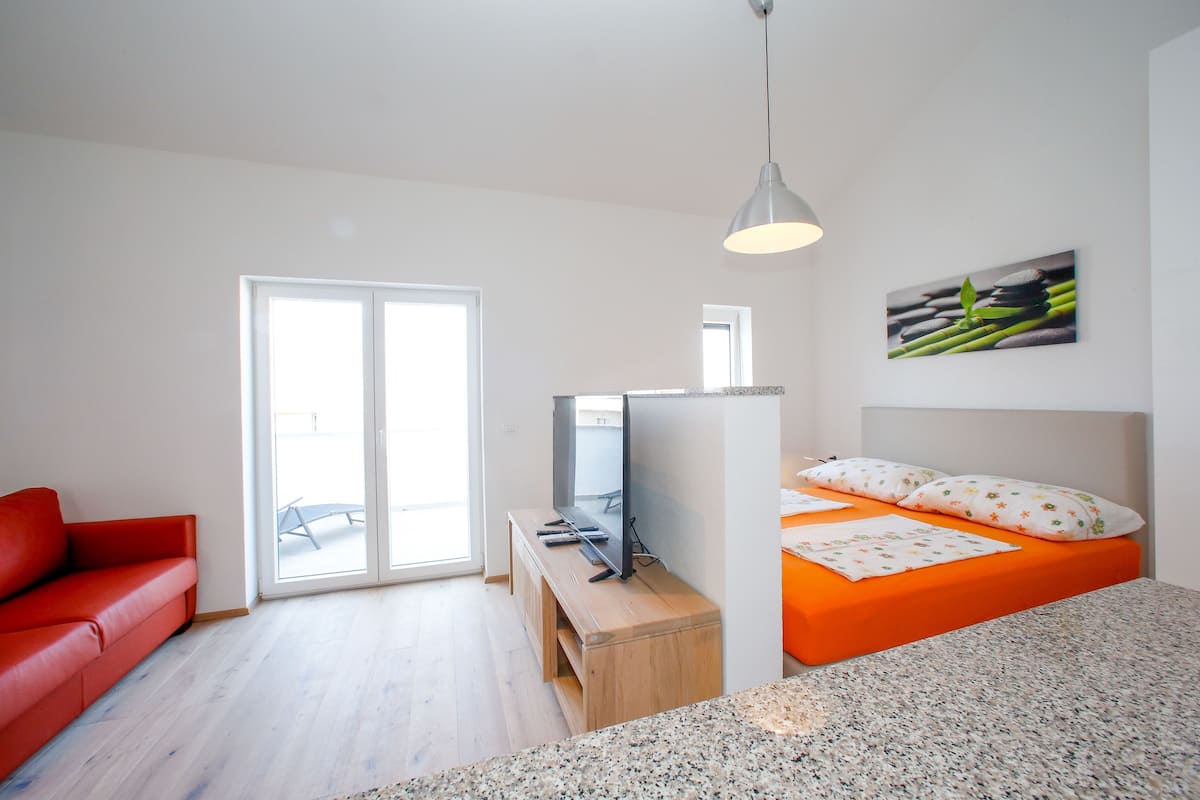 A modern room with a red sofa, a bed with orange bedding, a television on a wooden stand, and a painting on the wall.
