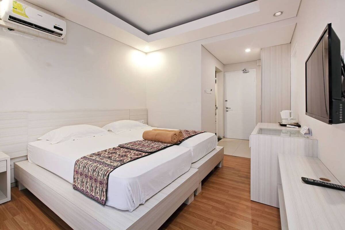 A hotel room with a bed, bedside table, TV, and an air conditioning unit.