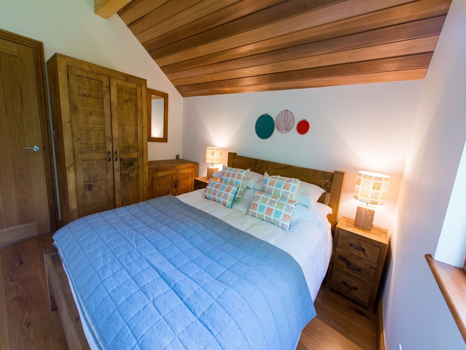 Un dormitorio con techo de madera, una cama grande con colcha azul, dos mesitas de noche con lámparas, un armario de madera y un espejo.
