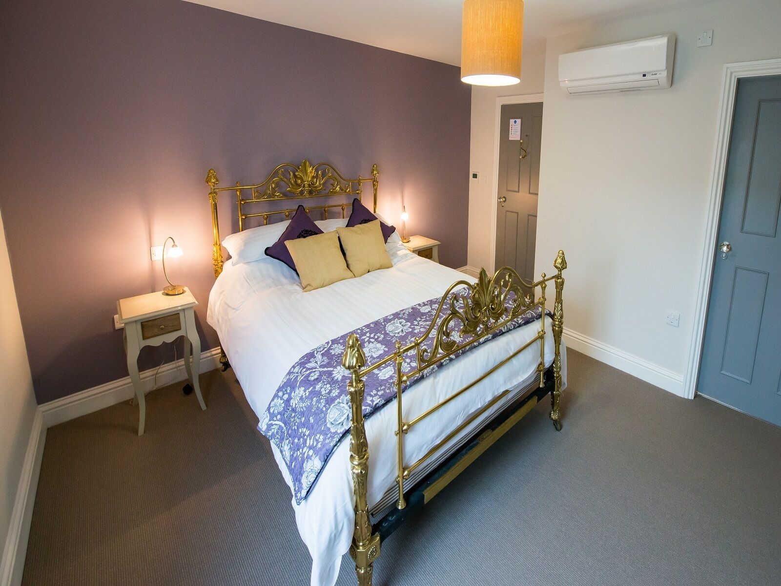 Una cama bien hecha con estructura dorada, ropa de cama blanca y almohadas moradas y beige. Se observa una mesita de noche con una lámpara, un aire acondicionado de pared y una puerta a la derecha.