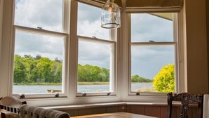 A dining area with a wooden table and chairs, large windows with a view of a lake and trees, and a hanging light fixture. - The Boathouse (Great Yarmouth)