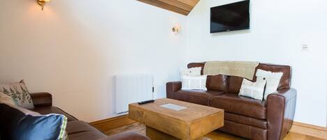 Una sala de estar con un sofá de cuero marrón, una mesa de centro de madera y un radiador blanco. Un televisor de pantalla plana está montado en la pared sobre el sofá.