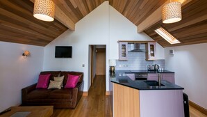 A cozy loft-style living space with a wooden ceiling, a brown leather sofa with colorful cushions, a small kitchen area with a black countertop, and pendant lighting. - The Boathouse (Great Yarmouth)