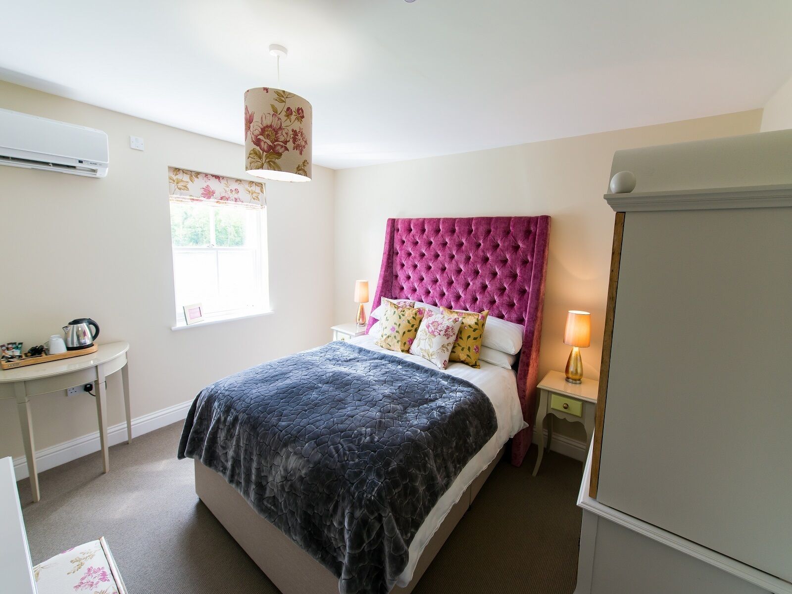 A bedroom with a bed, a bedside table, a lamp, a window, and an air conditioning unit.