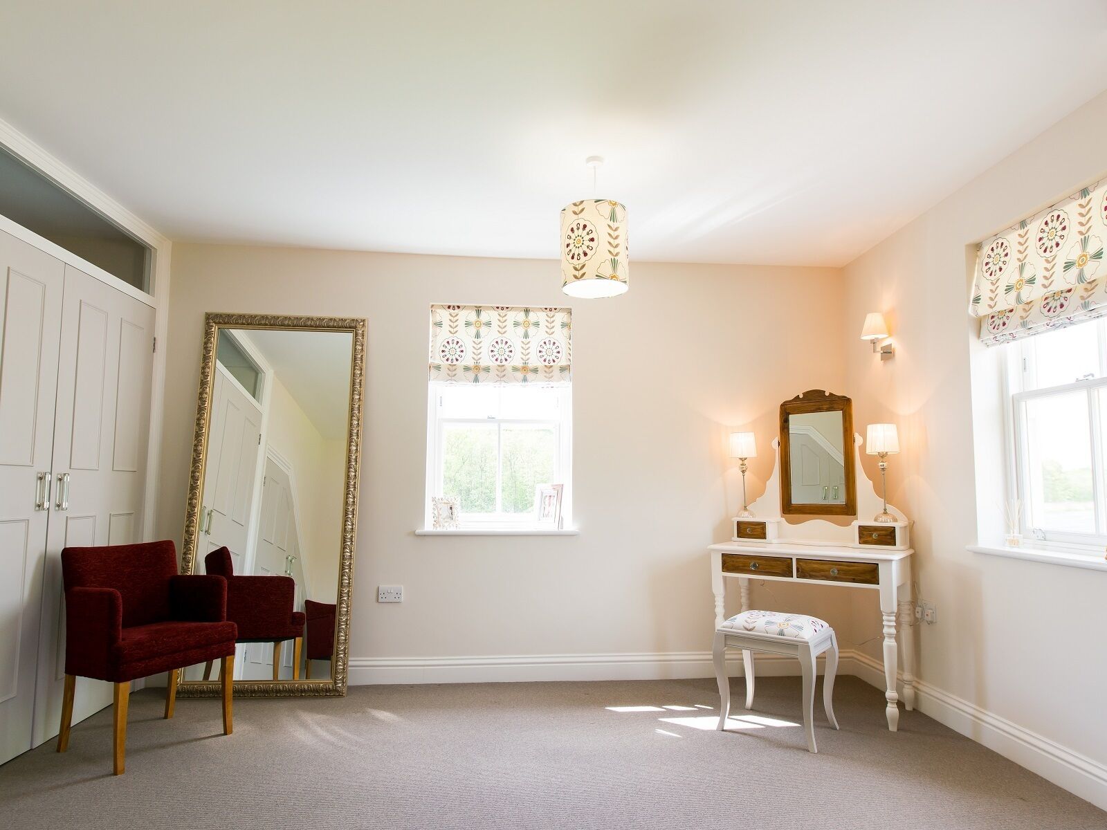 Une pièce avec un grand miroir, une coiffeuse avec un miroir, une chaise rouge, un banc blanc et une fenêtre ornée de rideaux à motifs.