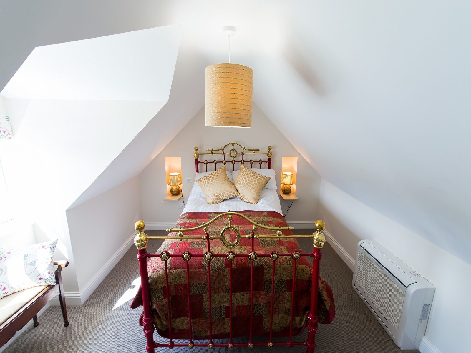 Un dormitorio con techo inclinado, cama con estructura metálica roja, ropa de cama estampada, dos lámparas de noche y un radiador.