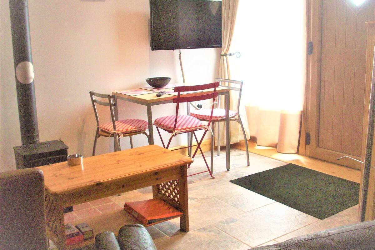 A small, cozy room with a wooden table and chairs, a television mounted on the wall, and a window with curtains.