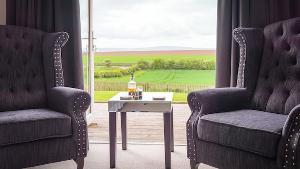 A room with two upholstered armchairs facing a table with a view of a green field and distant hills. - Garleton Lodge (Haddington)