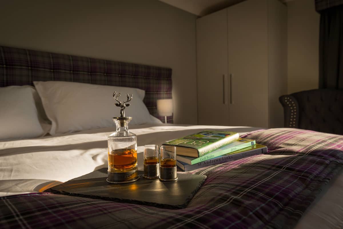 Una cama bien hecha con una colcha de cuadros, un libro, una botella y dos vasos para chupitos sobre una bandeja.
