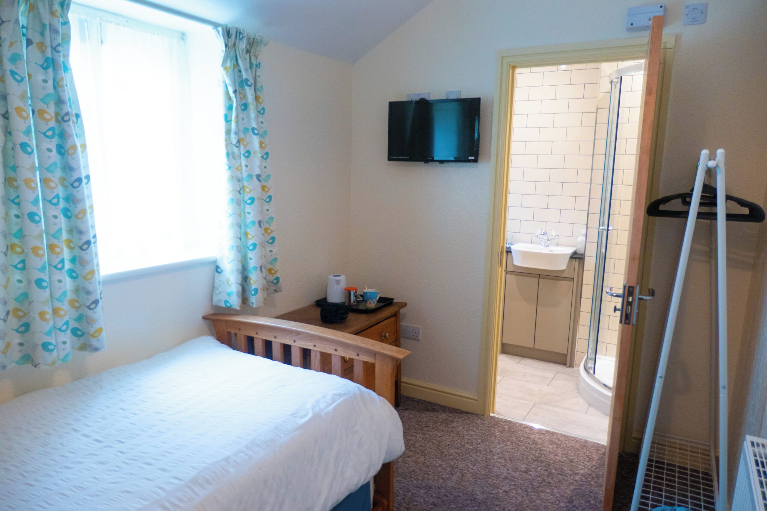 Un dormitorio con cama, televisor montado en la pared, una mesita con una taza y pañuelos, y un baño visible a través de una puerta abierta.