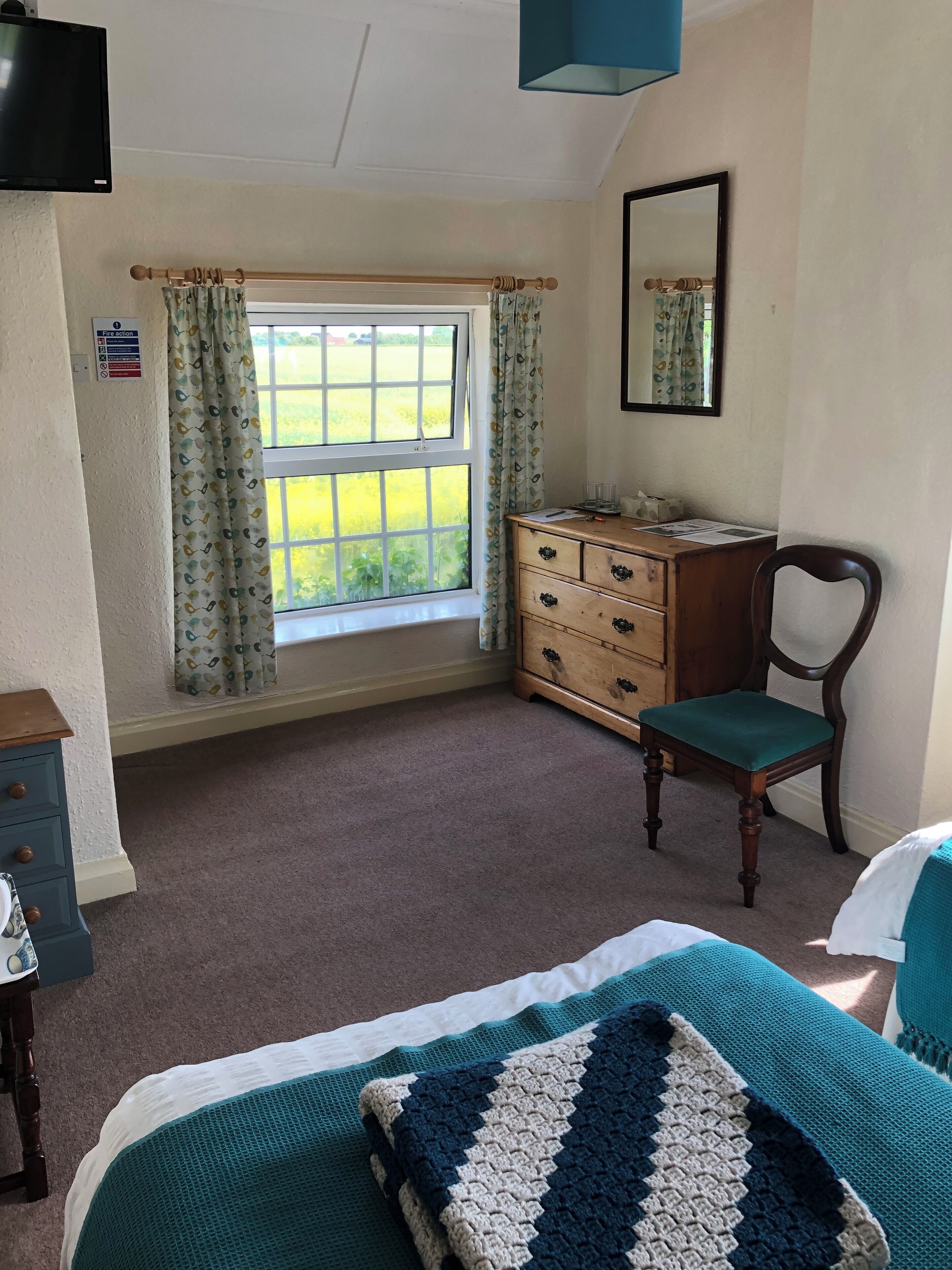 A bedroom with a bed, a wooden dresser, a chair, a window with curtains, a mirror, and a fire action sign.