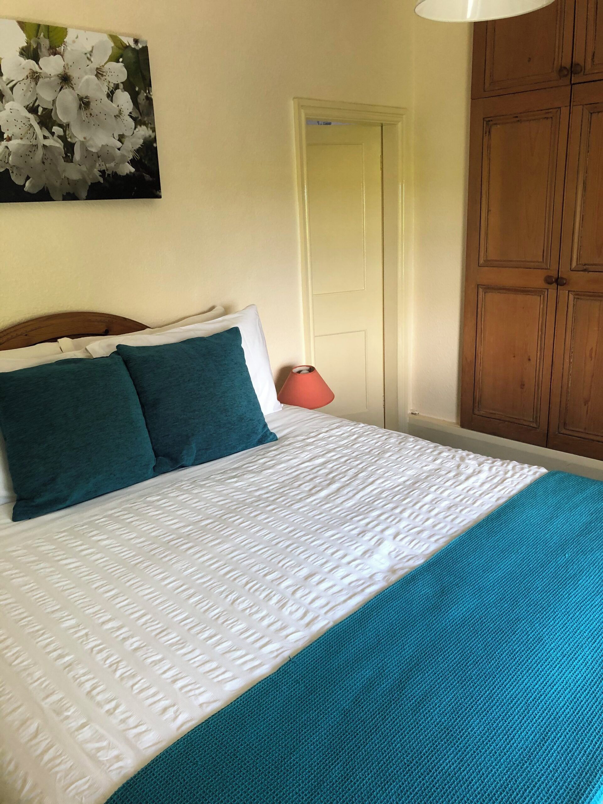 A neatly made bed with a white and teal bedspread, a wooden headboard, and a red bedside lamp.