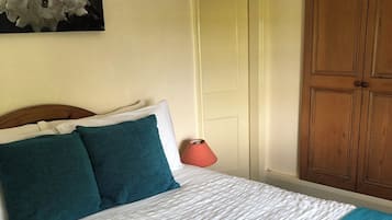 Uma cama bem arrumada com colcha branca e azul-turquesa, uma cabeceira de madeira e um abajur vermelho na mesa de cabeceira.