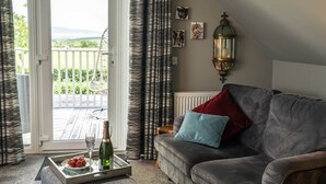 A living room with a grey sofa, a wooden coffee table with a bottle and a plate of food, a balcony with a view of greenery and mountains, and a wall-mounted lantern. - Dunroamin Apartment (Glasgow)