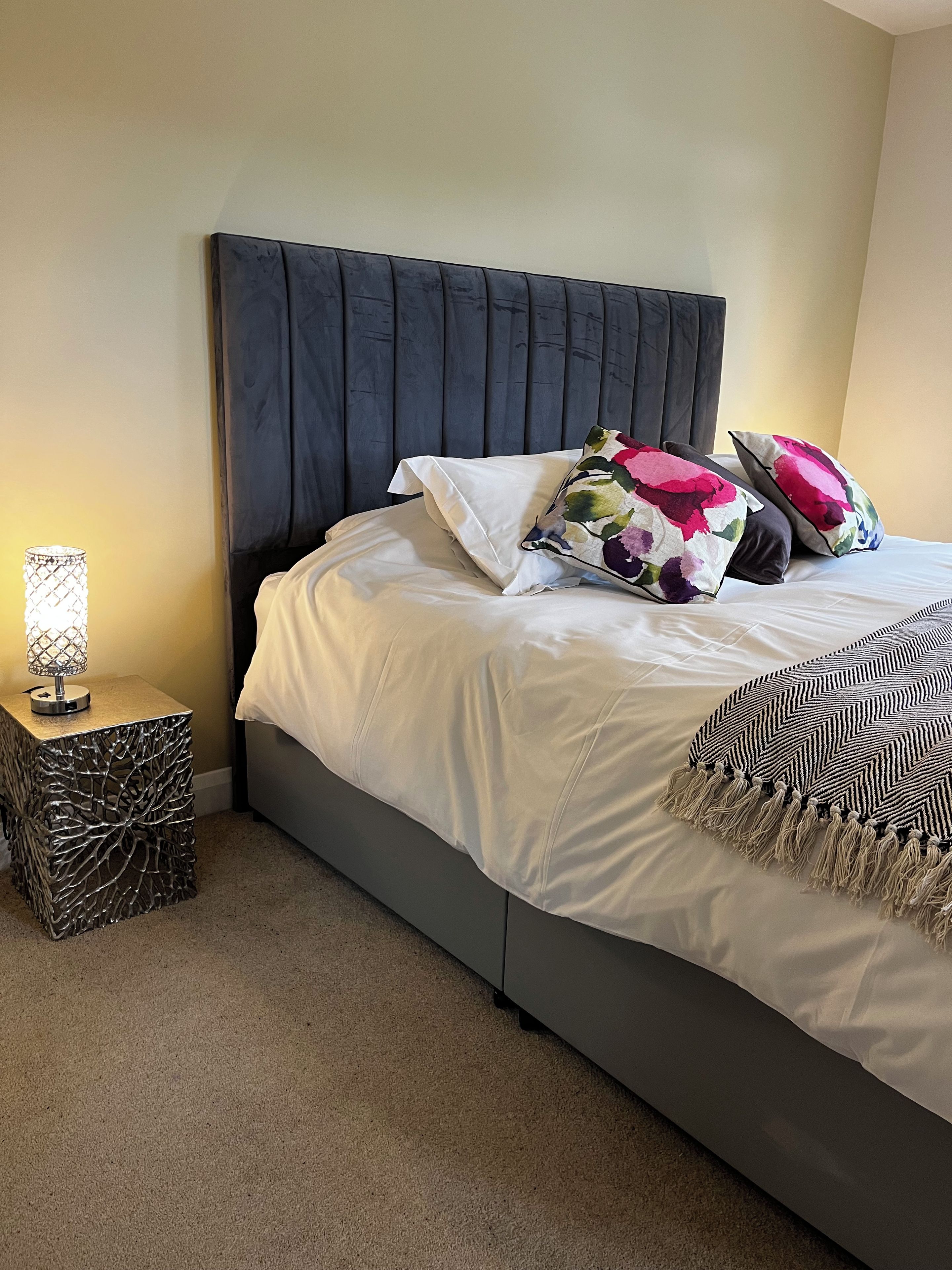 A bed with a dark headboard, white bedding, and decorative pillows. A small bedside table with a lamp and a decorative metal base is visible.