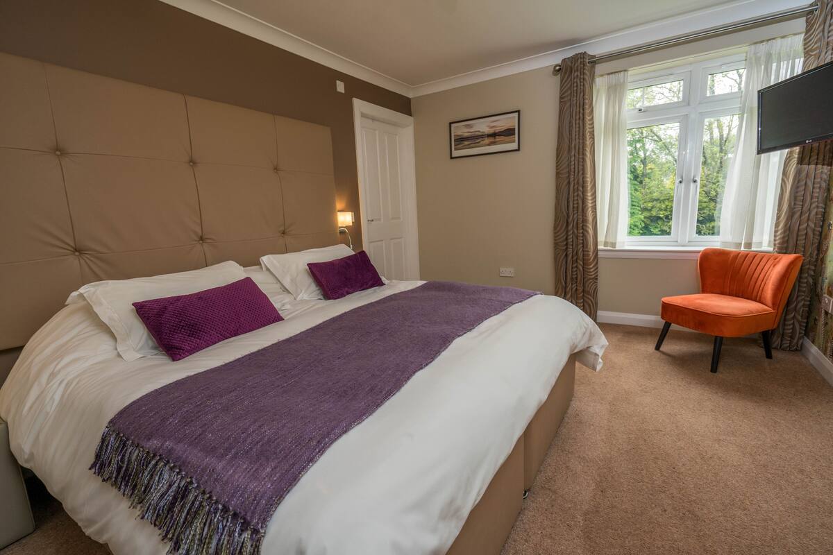 A bedroom with a large bed, a chair, a window with curtains, and a flat-screen TV mounted on the wall.