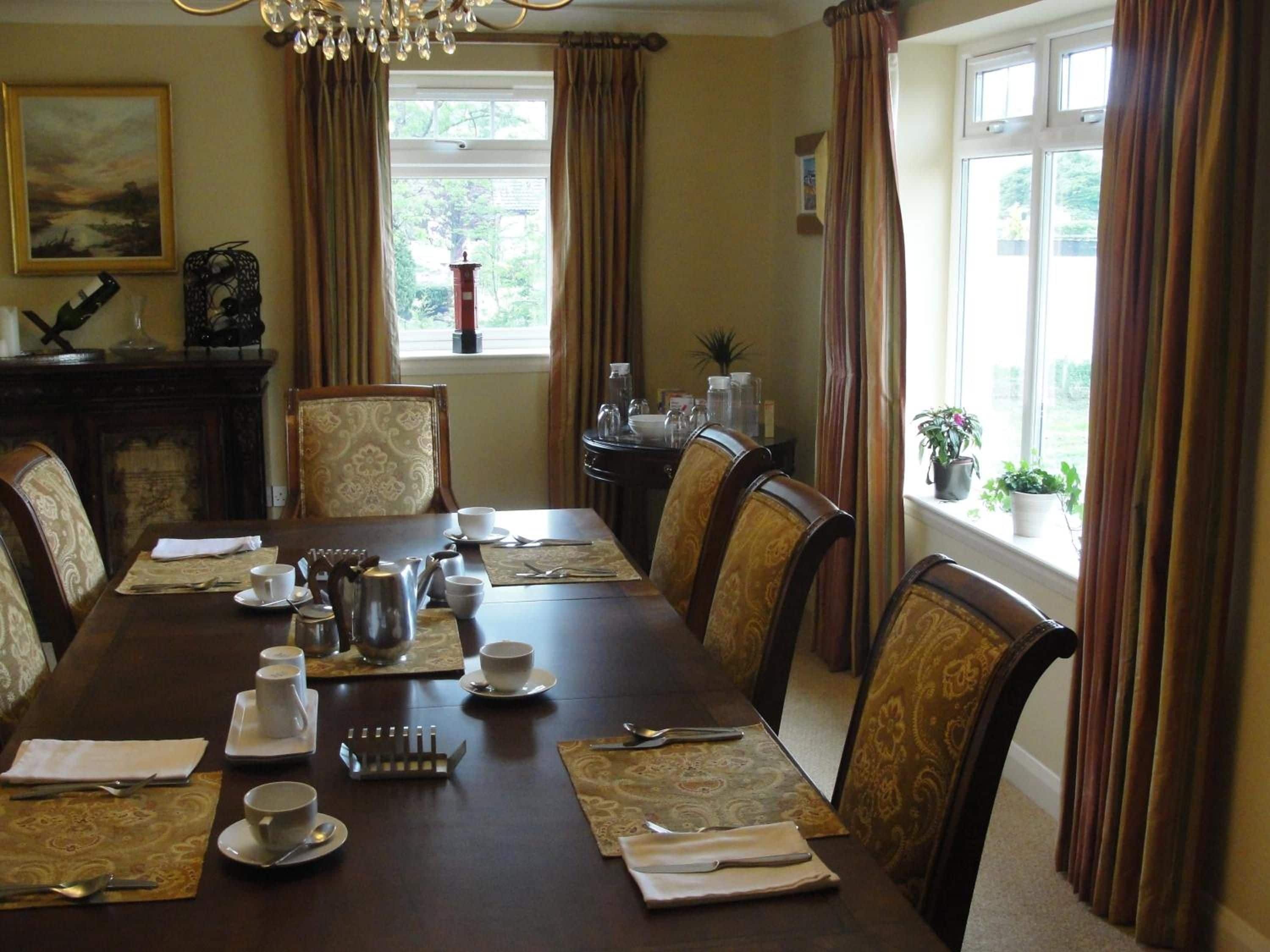 Une salle à manger élégante, avec une longue table dressée pour un repas, entourée de chaises, et une fenêtre agrémentée de rideaux.