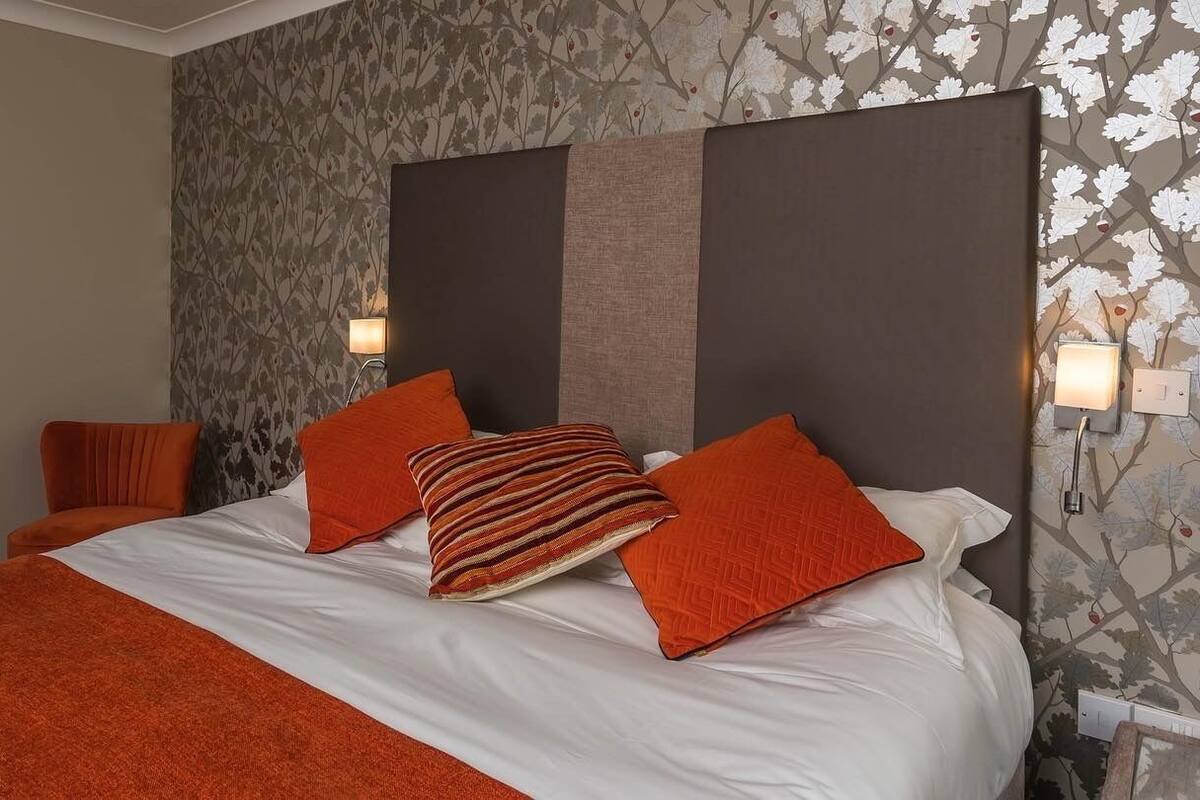A hotel room with a large bed, orange bedding, a patterned wallpaper, and a modern headboard.