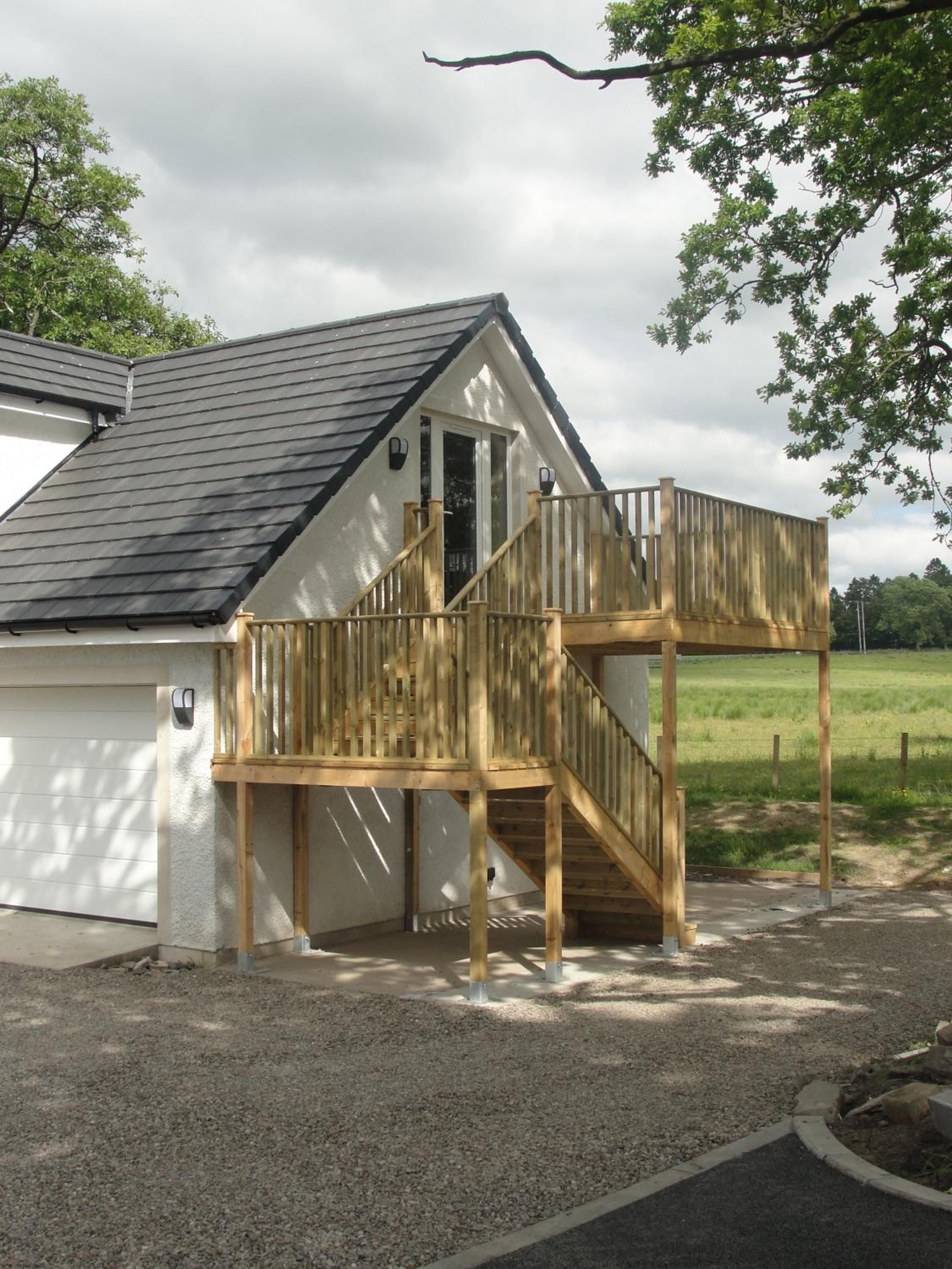 A two-story wooden deck attached to a white building with a dark roof, a garage door, and a gravel driveway.