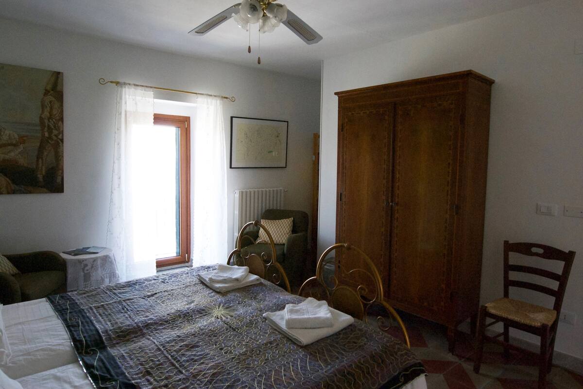 A bedroom with a bed, two chairs, a wooden wardrobe, a radiator, and a ceiling fan.
