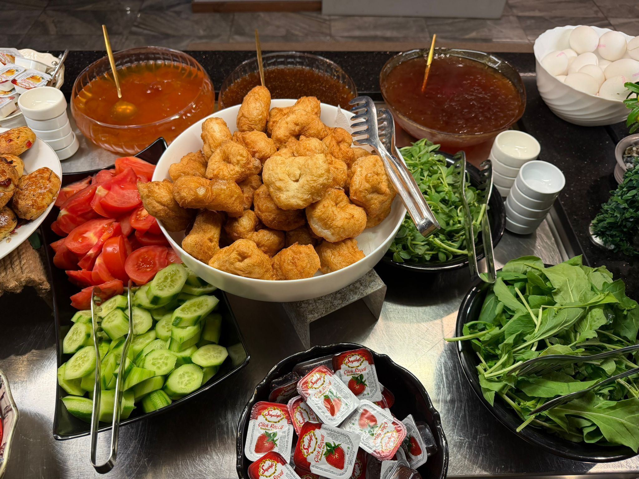A buffet table with various food items including fried chicken, sliced tomatoes, cucumbers, leafy greens, and packaged snacks.