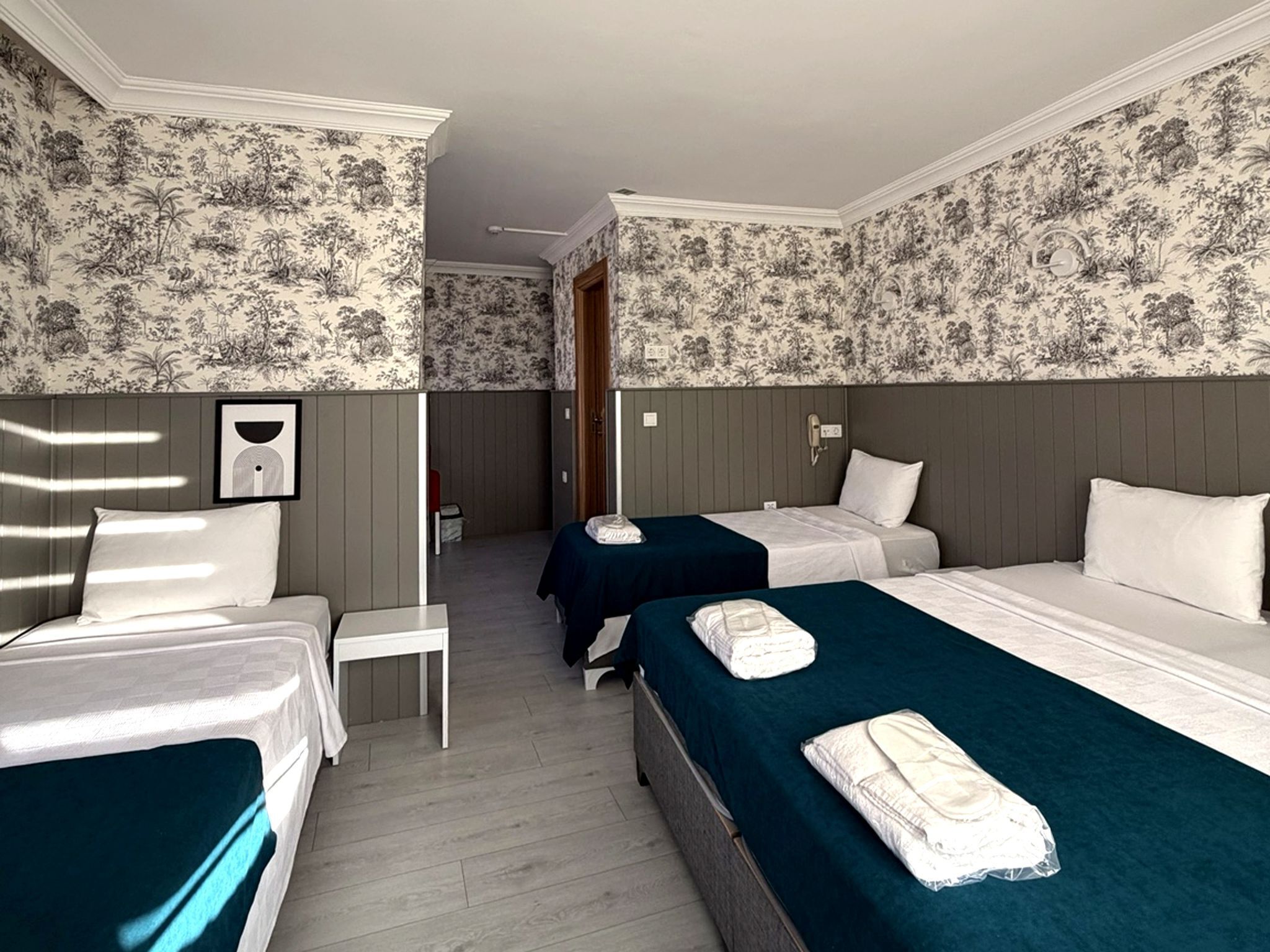 A hotel room with two single beds, a small desk, and a chair. The room has floral wallpaper and wooden paneling.