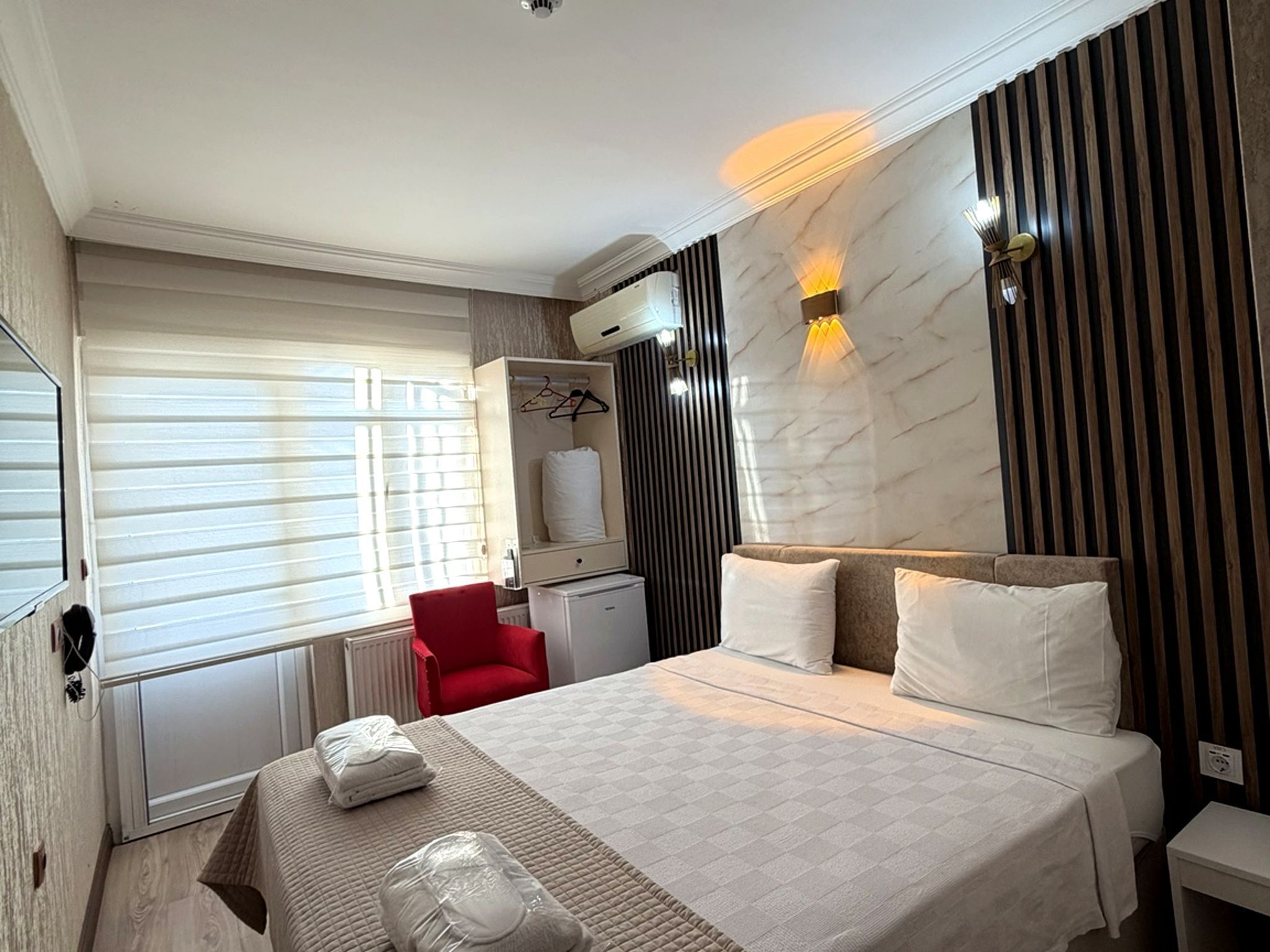 A hotel room with a bed, a red chair, a wardrobe, and a wall-mounted heater.