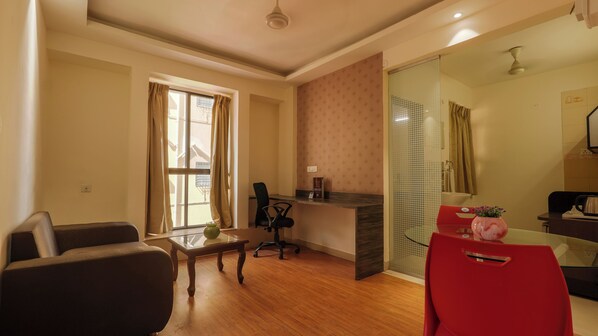 A modern living room with a wooden floor, a sofa, a dining table with red chairs, a small office desk, and a glass partition.