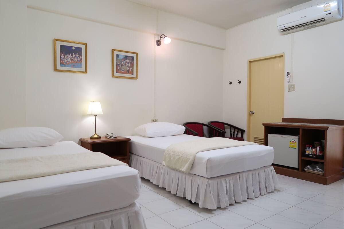 Quarto de hotel com duas camas, uma mesa de cabeceira com abajur, um pequeno frigorífico e ar condicionado fixado na parede.