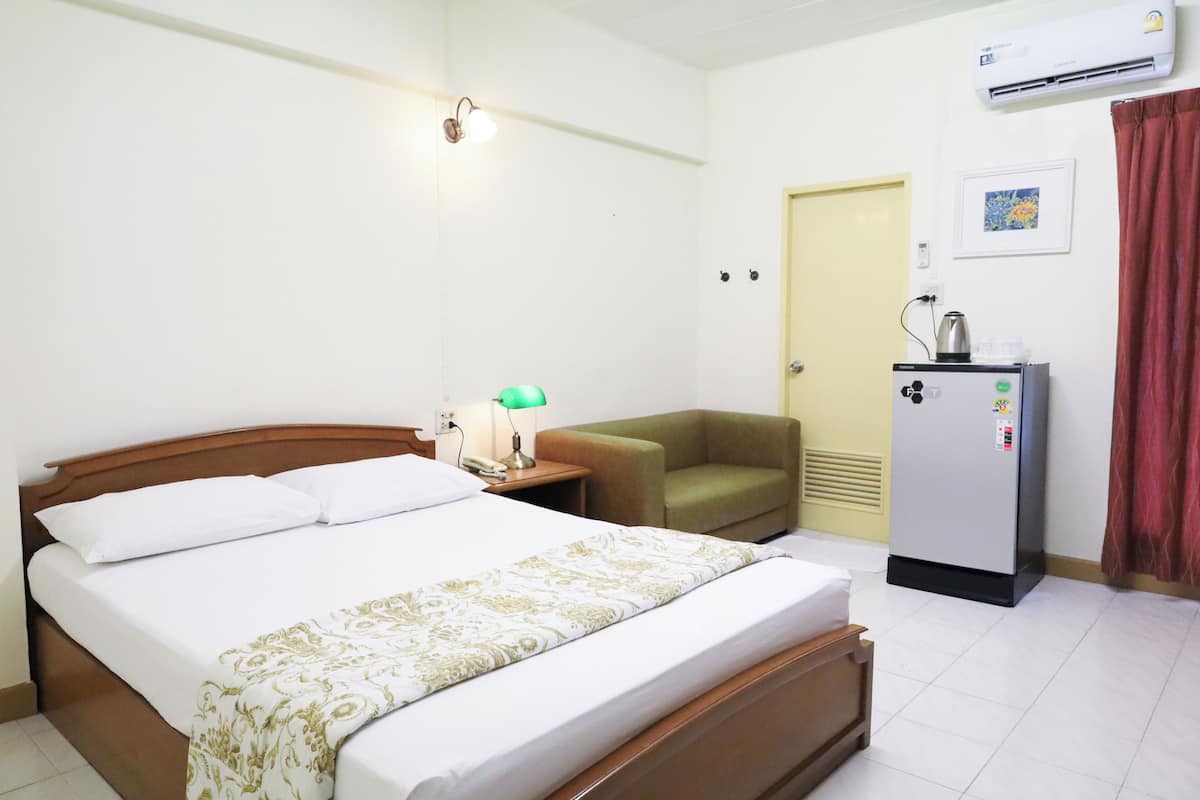 Quarto de hotel com cama, mesa de cabeceira, poltrona verde, frigorífico e porta.