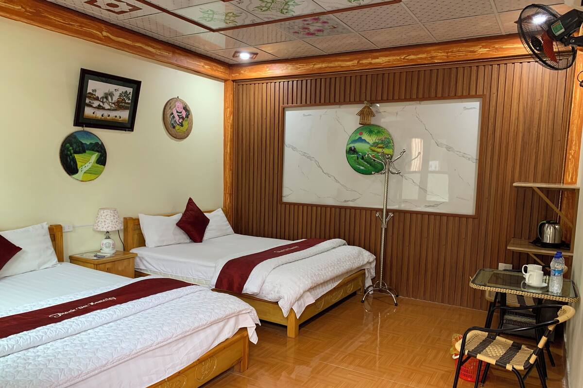 Une chambre d’hôtel avec deux lits, un plancher en bois, un ventilateur de plafond, un téléviseur fixé au mur et une petite table avec une chaise.