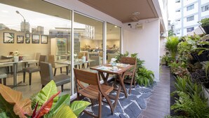 A modern outdoor dining area with wooden tables and chairs, surrounded by potted plants and a glass wall offering a view of the interior. - Bangkok Loft Inn (Bangkok)