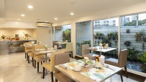 A modern dining area with multiple tables and chairs, a counter with food items, and large windows with a view of the city and plants. - Bangkok Loft Inn (Bangkok)