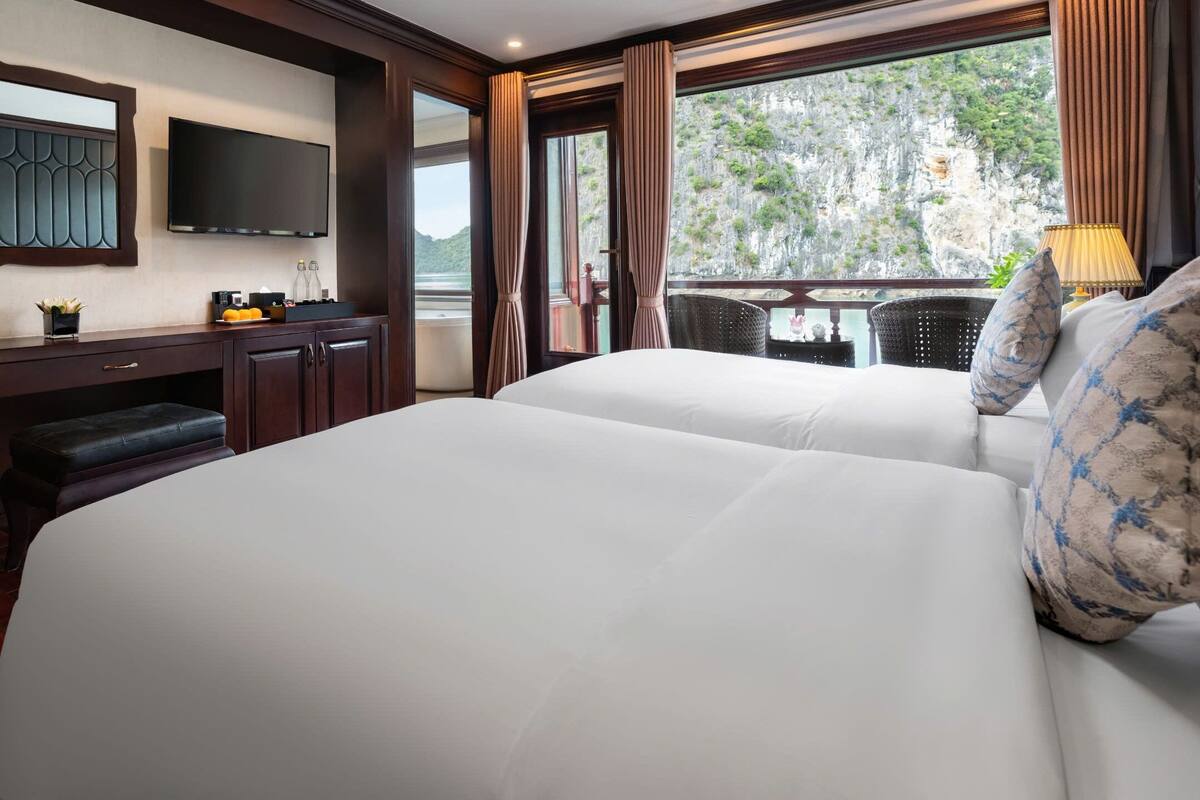 A hotel room with two beds, a flat-screen TV, a desk with a computer, and a window with a view of a mountainous landscape.