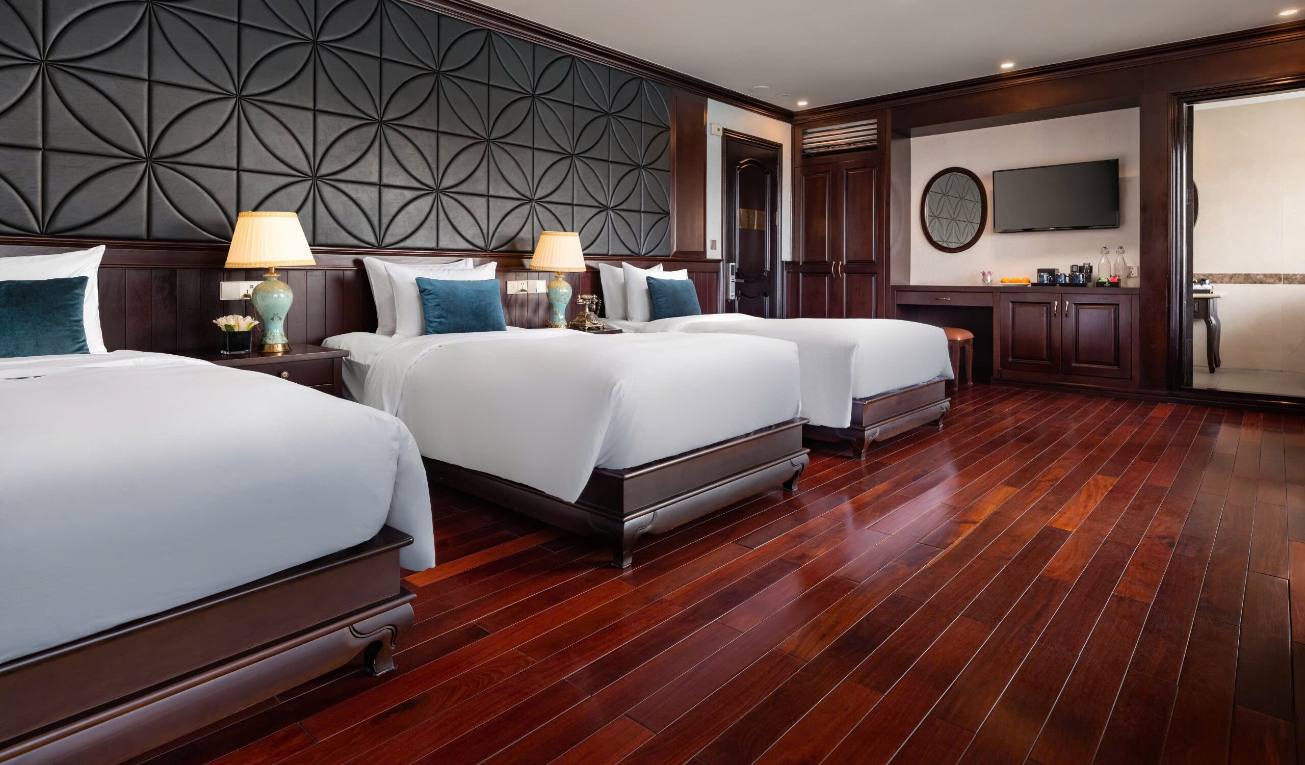 A hotel room with three beds, a dark wood-paneled headboard, a TV, and a kitchen area.