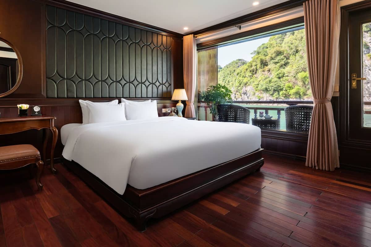 A large bed with a dark headboard, a bedside table with a lamp, a chair, and a view of a balcony with plants.