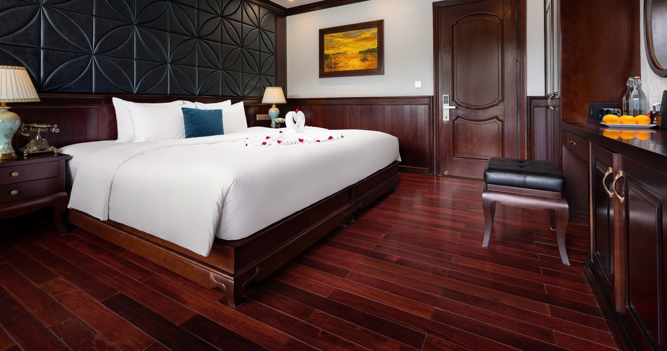 A hotel room with a large bed, dark wood paneling, a wooden floor, a desk with a lamp, and a door with a handle.