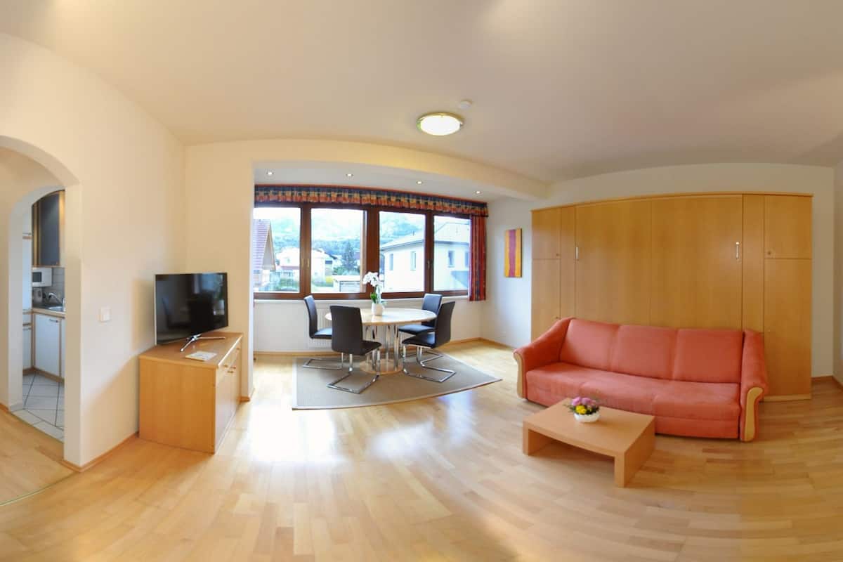 A modern living room with a sofa, coffee table, and dining area with chairs and a table. There is a kitchen area with a microwave and cabinets. The room has wooden flooring and large windows with curtains.