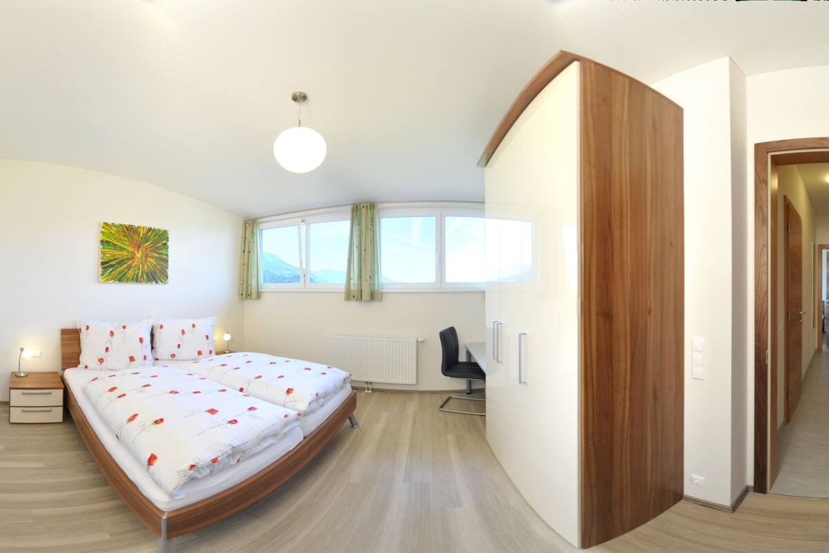 A modern bedroom with a curved bed, a bedside table, a chair, and a view of the outdoors through large windows.