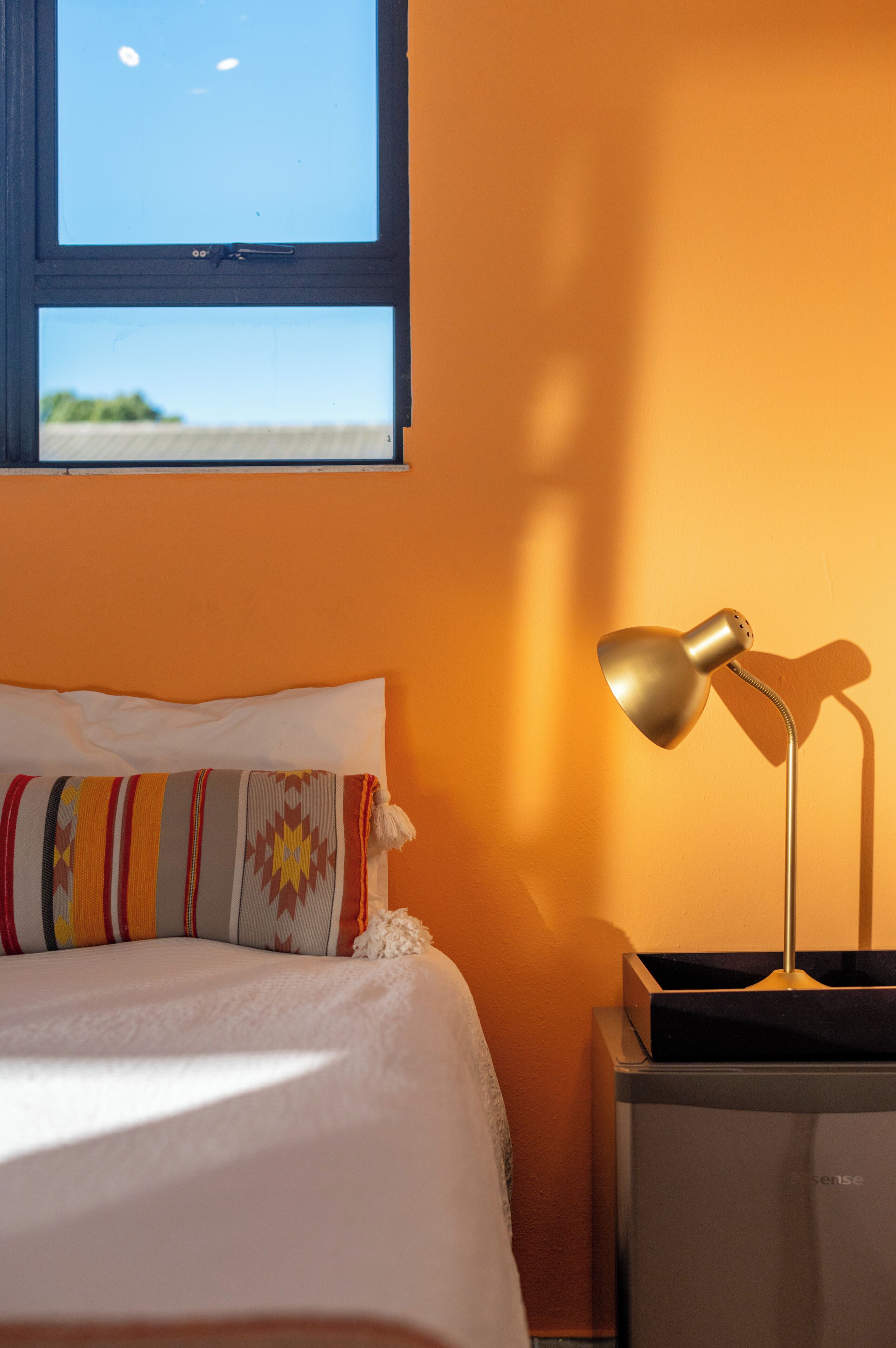 Una cama con un almohadón a rayas, una lámpara de noche y una ventana con vista al cielo.