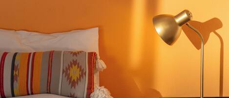 Una cama con un almohadón a rayas, una lámpara de noche y una ventana con vista al cielo.