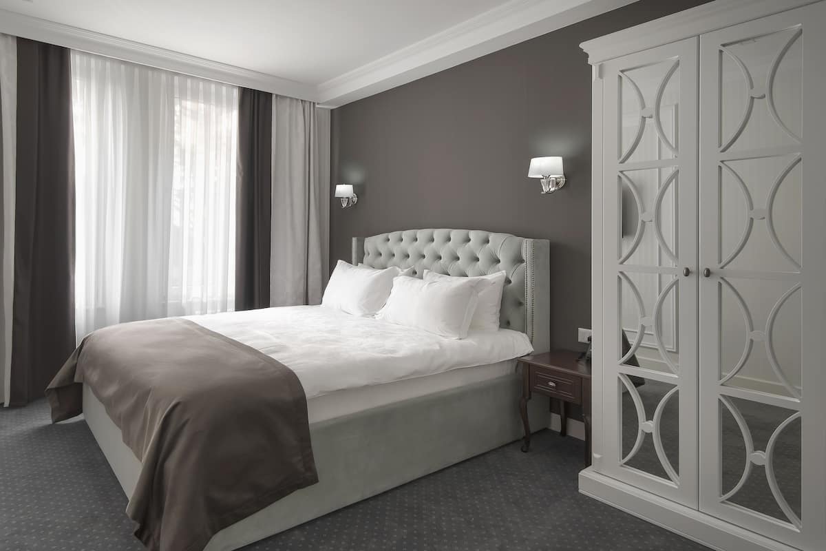 A neatly made bed with a grey headboard, a bedside table with a lamp, and a wardrobe with decorative panels.