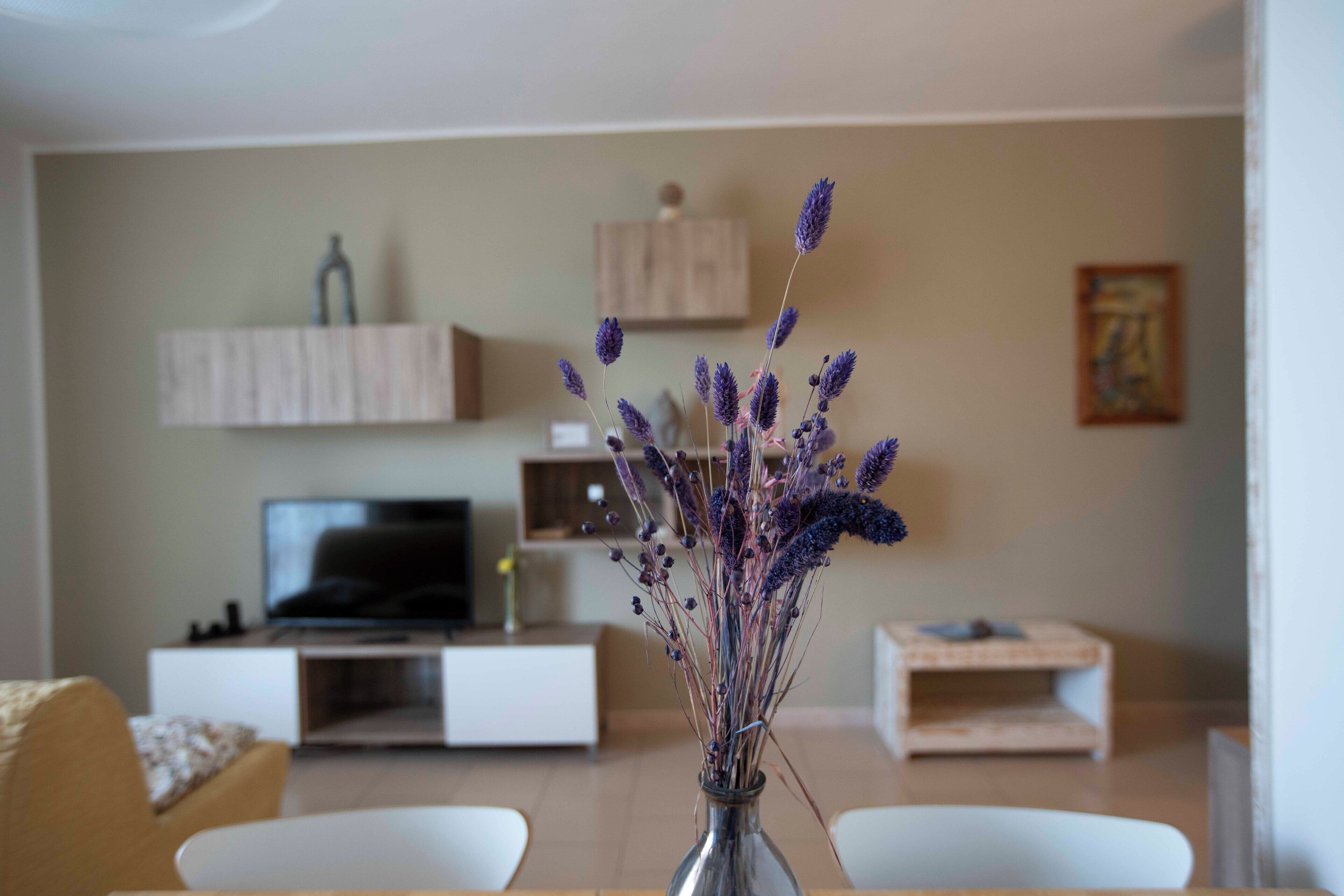 Un soggiorno con televisione, un vaso con fiori viola e un mobiletto in legno.